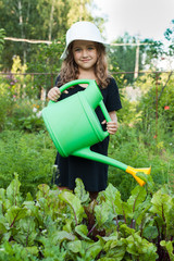Beautiful Little Girl Watering From A Watering Can In Her Hand On Vegetable Garden At Summer. Little Girl And Green Watering Can.