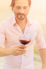 Confident and happy sommelier. Toned image of bearded mature man in pink shirt tasting red wine and looking at glass isolated on sea.