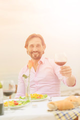Toned picture of handsome mature man smiling for the camera while holding glass of red wine in his hand. Handsome man in pink shirt resting in restaurant or cafe.