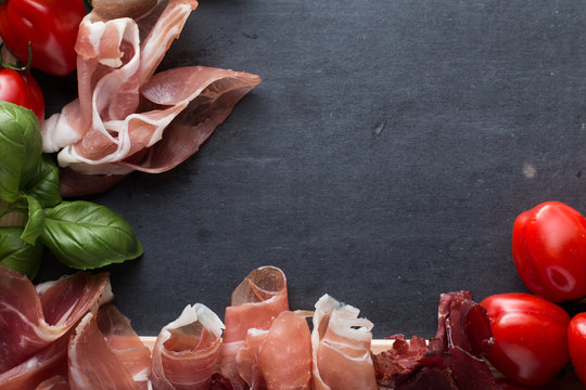 Jambon mix. Ham. Traditional Italian and Spanish salting, smoking, dry-cured dish - jamon Serrano and prosciutto crudo sliced with herbs and tomatoes on dark stone background. Copy space. Closeup. 
