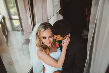 Fototapeta premium The charming bride and groom stand near window. Newlyweds near window embracing and kiss.