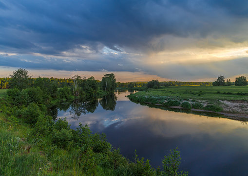 Summer evening, the river flows through a field, a forest in the distance, the sky dark blue clouds