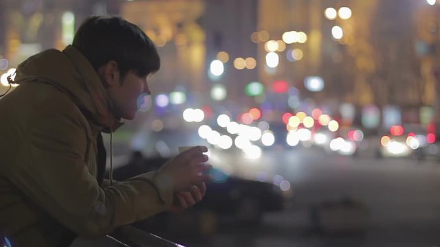 Young lonely man drinking hot tea on street, watching night city, sadness