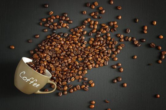 Fototapeta Overturn the mug with the inscription of coffee and scattered roasted coffee beans lying on a black wooden background. Flat lay. Top view. Close up.