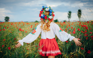 young beautiful woman with blond long hair in national dress red skirt and a white shirt with a wreath of flowers in a field of poppies and wheat