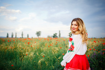 Obraz na płótnie Canvas young beautiful woman with blond long hair in national dress red skirt and a white shirt with a wreath of flowers in a field of poppies and wheat