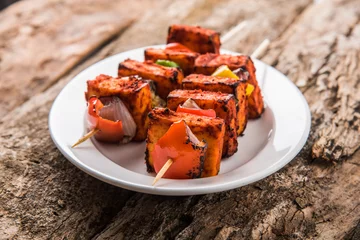 Fototapete Paneer Tikka Kabab - Tandoori Indian cheese skewers, malai paneer tikka / malai paneer kabab, chilli paneer served in white plate with barbecue stick and colourful capsicum and onion, with green sauce © StockImageFactory