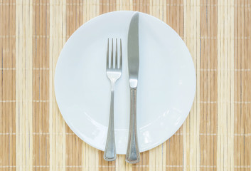 Closeup white ceramic dish with stainless fork and knife on wood mat textured background on dining table in top view in the food is very delicious concept