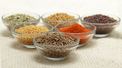 Different types of Indian spices in glass bowl, focus on cumin seeds on jute mat background. Front view.