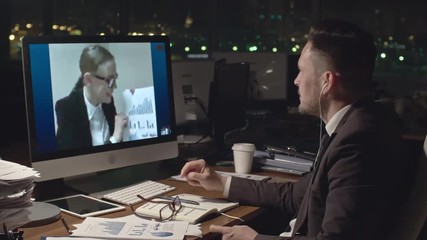 Medium shot of confident businessman sitting at desk in dark office and discussing documents with financial data with female business partner via video call on computer - Powered by Adobe
