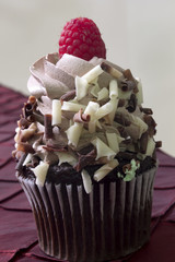 This is a photograph of a Chocolate cupcake topped with Chocolate frosting,Chocolate shavings, and Raspberry