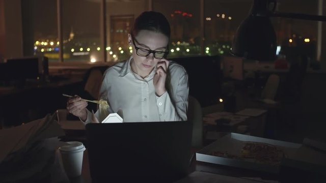 Young businesswoman sitting at desk in dark office, working on laptop, talking on mobile phone and eating noodles from box with sticks, medium shot on Sony NEX700 + Odyssey 7Q