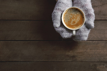 Female hands in knitted gloves holding cup of coffee on wooden background