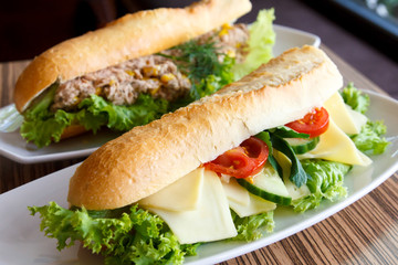 Cheese and salad baguette on white plate. Tuna baguette in backg