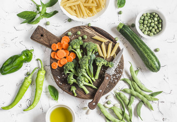 Raw ingredients for cooking vegetarian lunch - dry pasta, fresh broccoli, zucchini, green peas and beans, carrots, olive oil. On a light background, top view