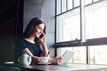 Young attractive girl with cute smile talking on mobile phone while sitting alone in coffee shop during free time and working on tablet computer. Happy female having rest in cafe. Lifestyle