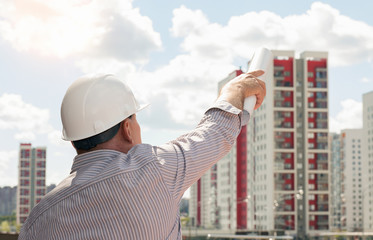 An engineer in white helmet pointing with blueprints in his hand on buildings on a background