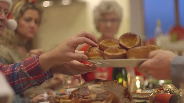 A family enjoy their Christmas dinner at the table together. One of the family members is passing around a plate of Yorkshire puddings for everyone to share.
