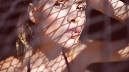 Closeup portrait of beautiful blonde woman wearing black swimwear sitting on a rooftop in a sunny summer day with shadow net reflection from metal sunshade on her face over black wall