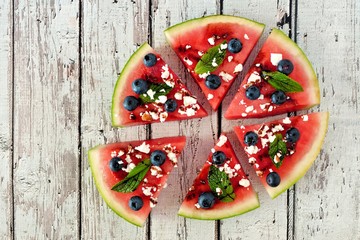 Fototapeta na wymiar Watermelon pizza in slices with blueberries, mint, balsamic glaze and feta, above view on rustic white wood