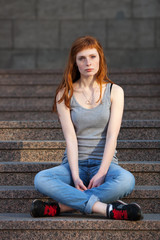 Fototapeta na wymiar Young redhead caucasian woman in grey shirt blue jeans black sneakers sitting on stairs outdoor portrait