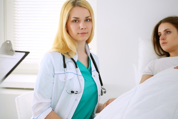 Young beautiful female doctor sitting near the bed of her patient in hospital.