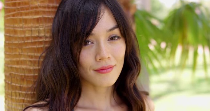 Smiling young woman leaning against a tropical palm tree as she cools off in the shade from the hot summer sun  closeup head and shoulders