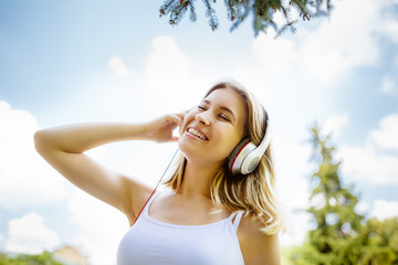 Teenage girl with headphones  outdoors on sunny summer day smiling. Beautiful millennial young woman listening to music relaxing. Horizontal, retouched,