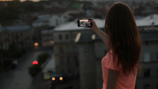 Young pretty woman with long hair taking photo, filming with cell phone at the top of building with city landscape at sunset. View from behind