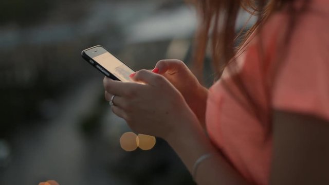 Closeup of young female hands scrolling pictures on phone, city lights bokeh on background