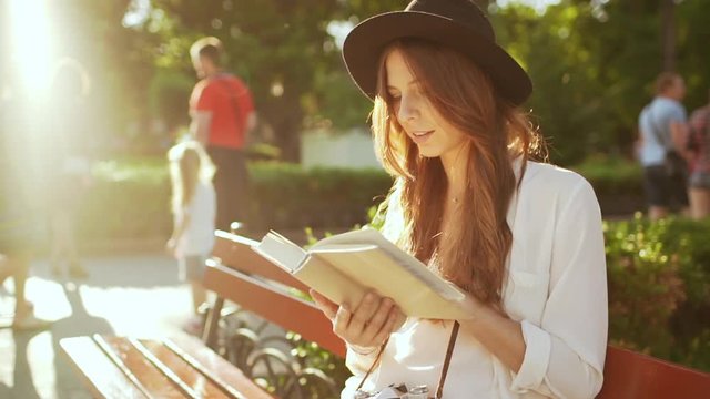 Young beautiful girl in hat reading book in city park. Slow motion.