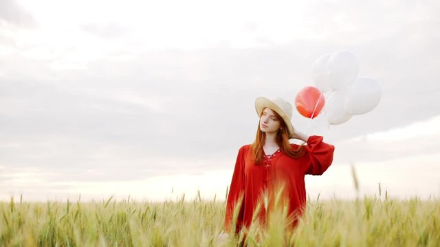 Young beautiful girl with foxy hair in red dress and hat with baloons walking in field. Slow motion.
