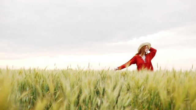 Young beautiful girl with foxy hair in red dress and hat walking in field. Slow motion.