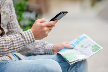 Closeup of male hands holding cellphone and city map outdoors on the street. Man using mobile smartphone to find famous attraction.