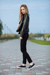 Happy young beautiful woman in black leather jacket black jeans slip-on posing for model tests in the summer park