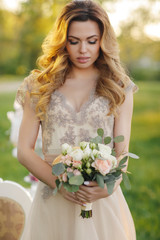 Young beautiful bride,a blonde woman with long curly hair,beautiful wedding dress beige in the hands holding wedding bouquet of the bride,posing standing in the summer Park near the white table