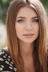 Naklejka premium Young woman brunette with long straight hair and gray eyes,long black eyelashes,a cute smile,dressed in a black summer dress with white daisies,posing on summer nature background outdoors