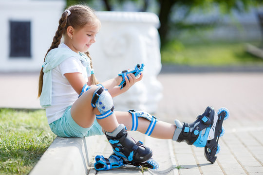 Blonde little girl with two long pigtails wearing a white t-shirt and blue shorts,wearing knee pads and protection on the elbows blue,spends time in a city Park,roller skating in the summer