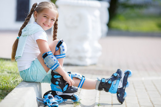 Blonde little girl with two long pigtails wearing a white t-shirt and blue shorts,wearing knee pads and protection on the elbows blue,spends time in a city Park,roller skating in the summer