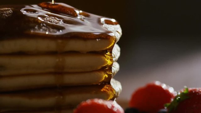 Maple syrup being poured over warm stacked pancakes with fruit, in slow motion