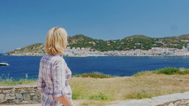 Steadicam shot: A female tourist walks along the coast, on the background of a Mediterranean village-resort of Cadaques in Spain