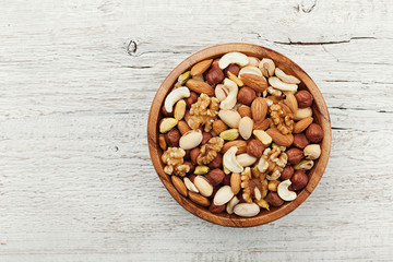 Wooden bowl with mixed nuts on white table from above. Healthy food and snack. Walnut, pistachios, almonds, hazelnuts and cashews. - obrazy, fototapety, plakaty