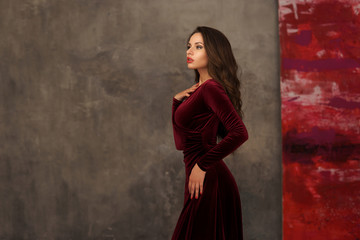 Fototapeta na wymiar Young beautiful and pretty elegant woman in long cherry red evening dress posing against gray concrete wall. Portrait of smiling happy cheerful girl