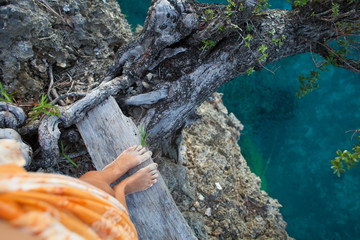 Highly Detailed Photo Woman Legs. Girl Looking Down Cliff Old Tree. Blue Ocean Water Blurred Background.Horizontal picture. Top View Image.