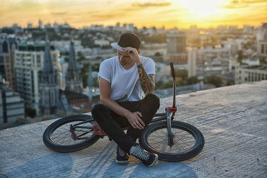 Young handsome hipster man sitting on the roof and posing on bicycle on sunset