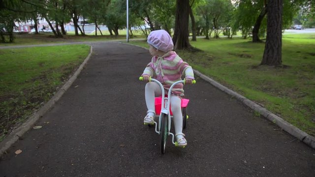 Little girl in striped sweater rides on a pink tricycle on the track of the city Park