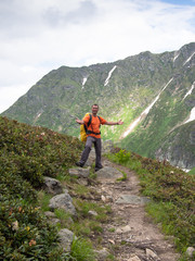 Adult male with a backpack standing on a footpath his arms outstretched on the background of snowy mountains with blue sky