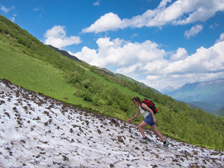 Young woman with a backpack and shorts up the hill through the snow against the blue sky with clouds