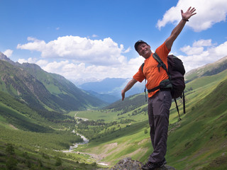Adult male with a backpack standing on a rock his arms outstretched on the background of snowy mountains with blue sky