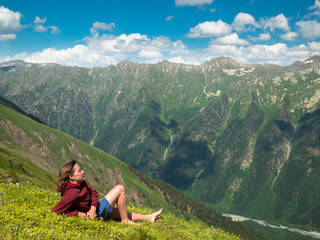 Fototapeta na wymiar Young woman lying in a meadow with flowers in front of the North Caucasus mountain range with the sky and clouds